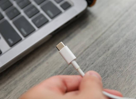 A close-up image of a USB-C cable with its connector in sharp focus, lying against a blurred background featuring a laptop on a wooden surface.